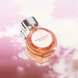 Paweł Retkowski Studio - Flakon perfum Mademoiselle Rochas na tle rozmytego, różowego nieba z chmurami, z widocznym srebrnym korkiem i delikatnym, pomarańczowym odcieniem płynu wewnątrz.