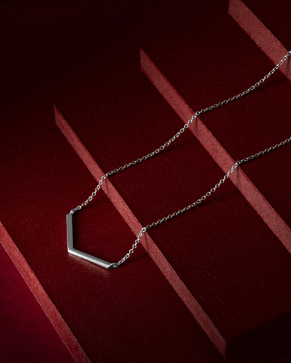 Srebrny naszyjnik z geometryczną zawieszką na tle stopniowanej, bordowej powierzchni.