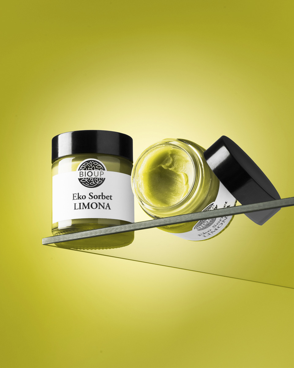 Dwa słoiczki z żółtym kremem Eko Sorbet Limona marki BIOUP, jeden stojący, drugi przechylony z otwartym wieczkiem, na szklanej półce na tle jednolitego, żółtego tła.