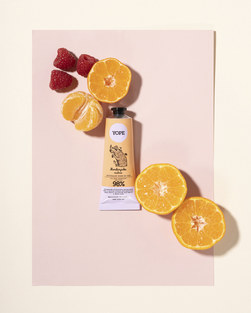 Krem do rąk YOPE o zapachu mandarynki i maliny, otoczony połówkami mandarynek i malinami na różowym tle.