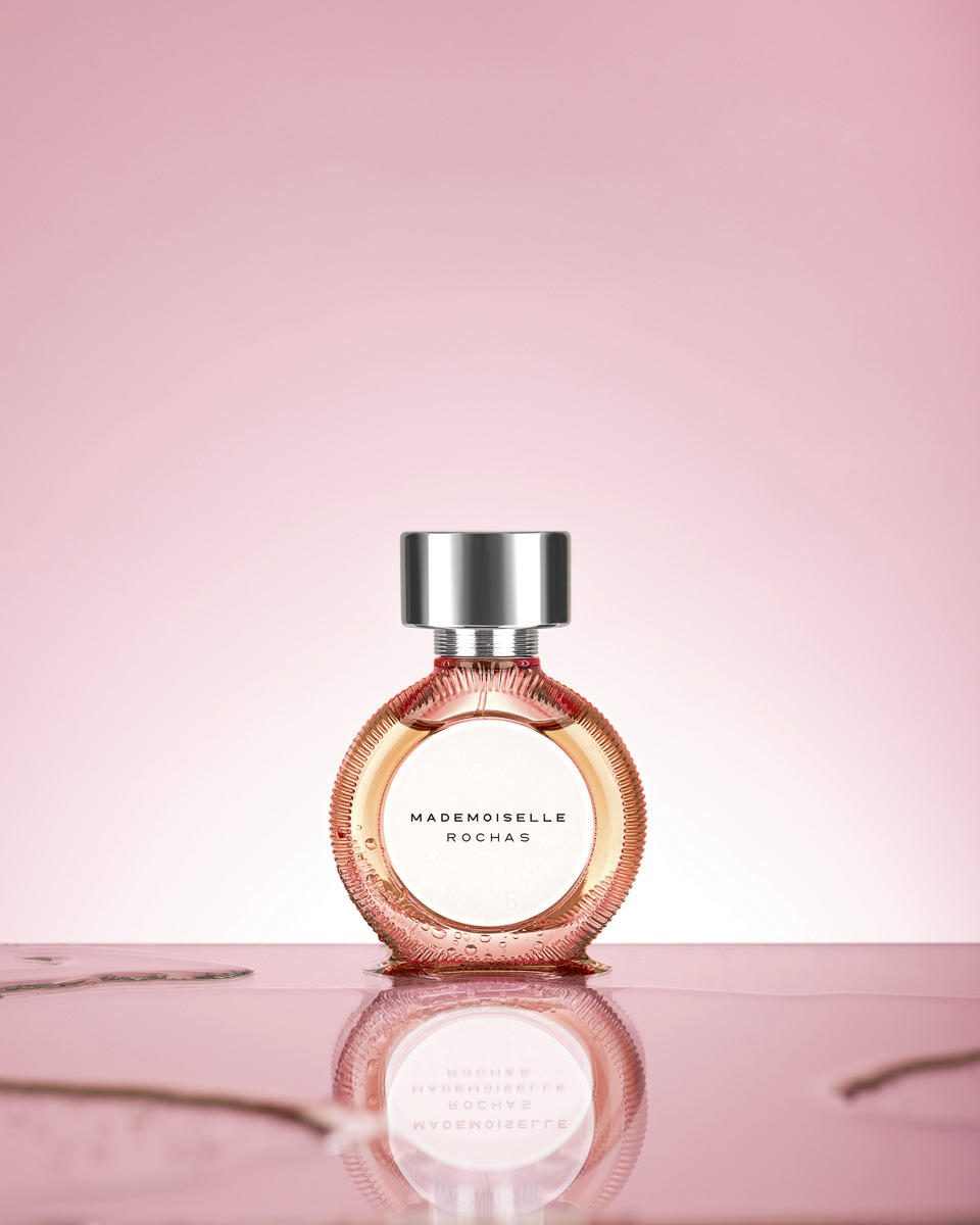 Butelka perfum Mademoiselle Rochas na błyszczącej powierzchni z odbiciem, na tle delikatnego różu.