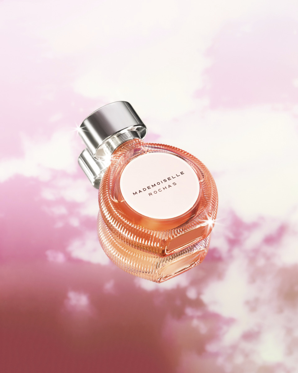 Flakon perfum Mademoiselle Rochas na tle rozmytego, różowego nieba z chmurami, z widocznym srebrnym korkiem i delikatnym, pomarańczowym odcieniem płynu wewnątrz.