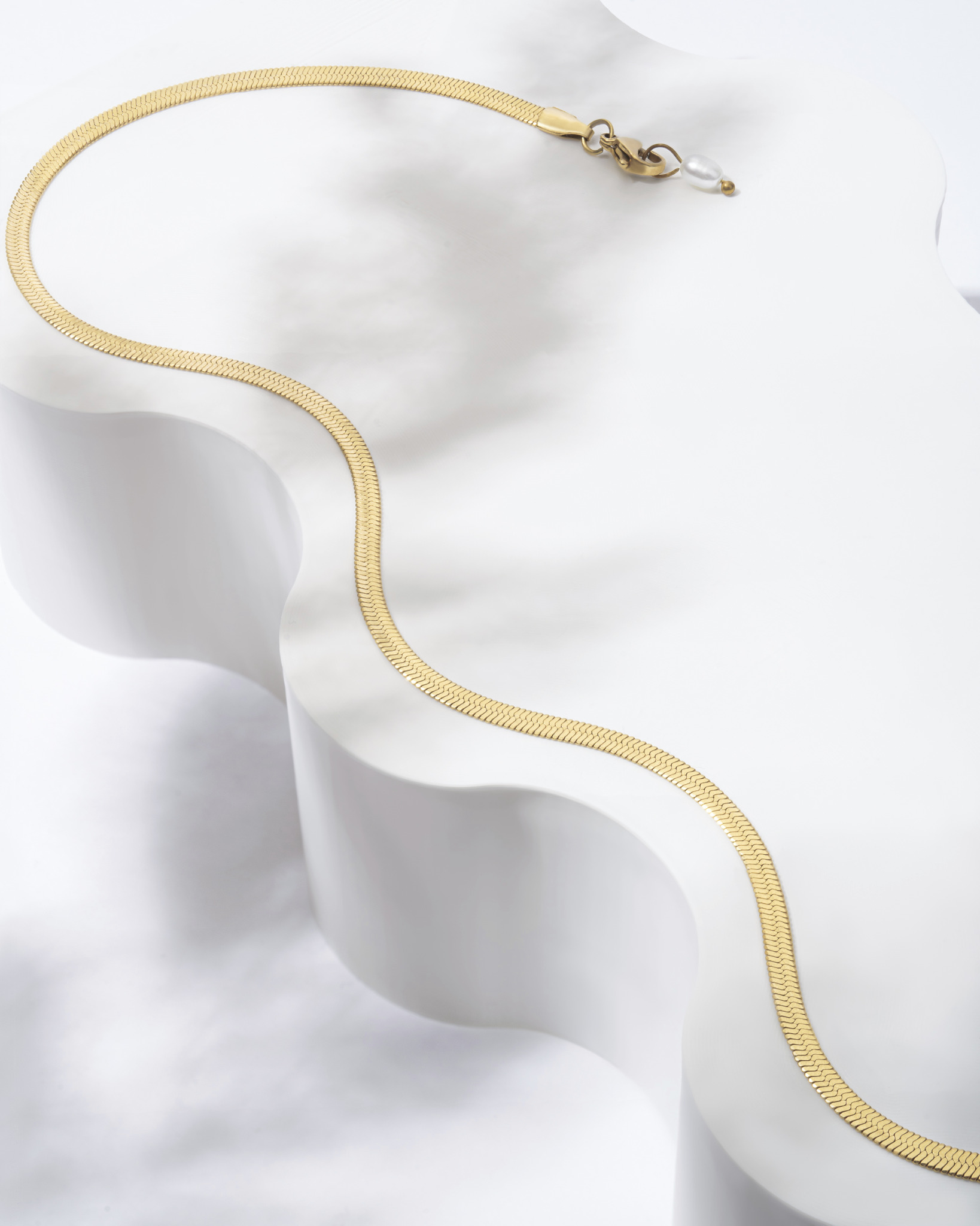 Złoty łańcuszek typu snake na białym, falistym postumencie, z zapięciem typu karabińczyk i perłą słodkowodną przy zapięciu.