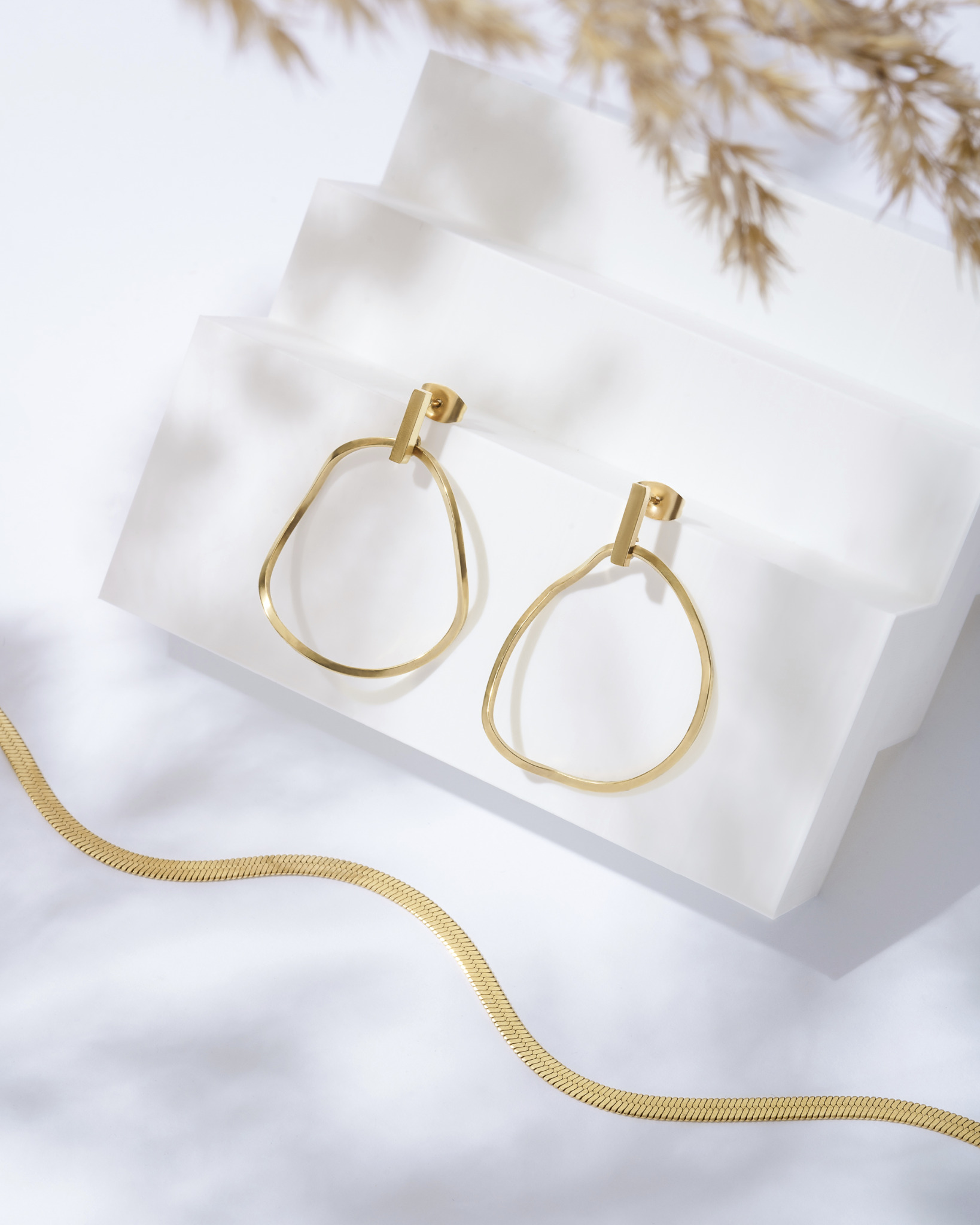 Dwa złote kolczyki o nieregularnym kształcie wiszą na białych, schodkowych blokach. Złoty łańcuszek leży obok, a w tle widać delikatne źdźbła trawy.