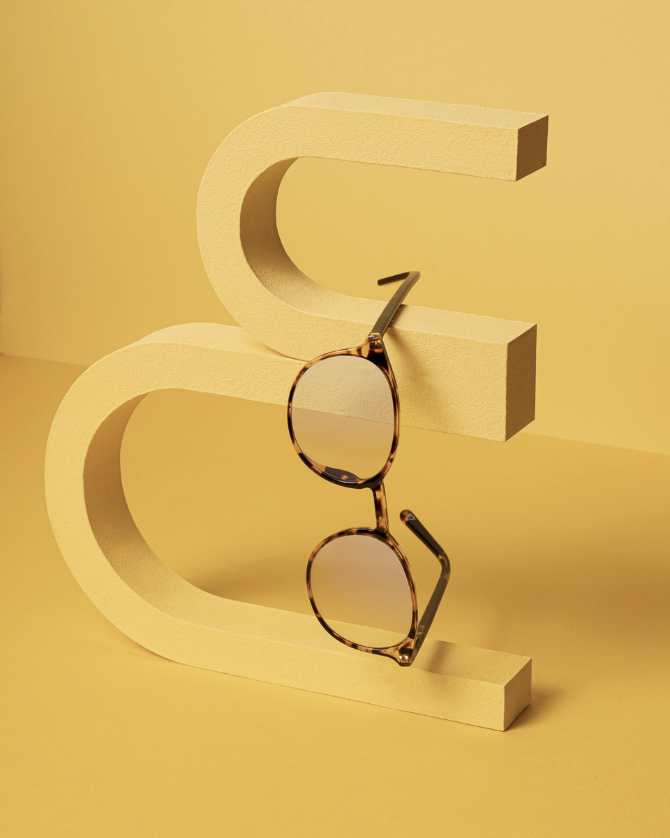 Okulary korekcyjne w brązowej oprawce w panterkę, zawieszone na żółtej, geometrycznej konstrukcji w kształcie litery 'S', na żółtym tle.