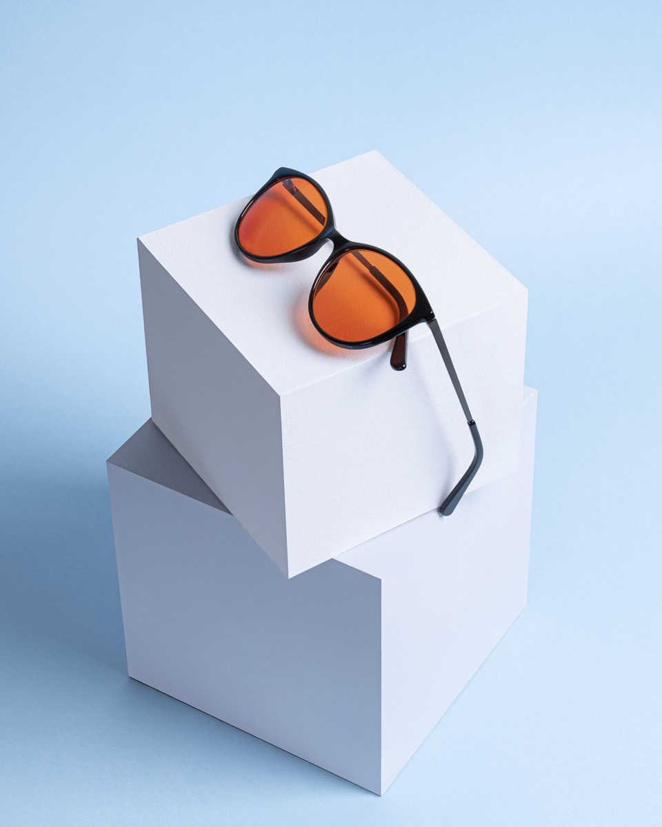 Czarne okulary z pomarańczowymi szkłami spoczywają na dwóch białych kostkach, na jasnoniebieskim tle.