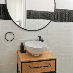 Nowoczesna łazienka z okrągłym lustrem w czarnej ramie, białą umywalką nablatową na drewnianej szafce z czarnymi elementami metalowymi, białe płytki ścienne i czarno-biała podłoga w kwiatowe wzory.