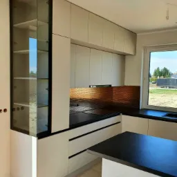 Nowoczesna kuchnia z białymi szafkami, czarnym blatem, miedzianym panelem ściennym z motywem plastra miodu i szklaną witryną po lewej stronie, widok na zewnątrz przez okno.