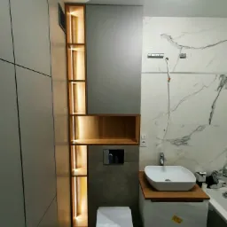 Nowoczesna łazienka w trakcie wykańczania: szara zabudowa meblowa z podświetlanymi półkami z drewna, wisząca toaleta, umywalka nablatowa na drewnianym blacie, marmurowe płytki częściowo zamontowane.