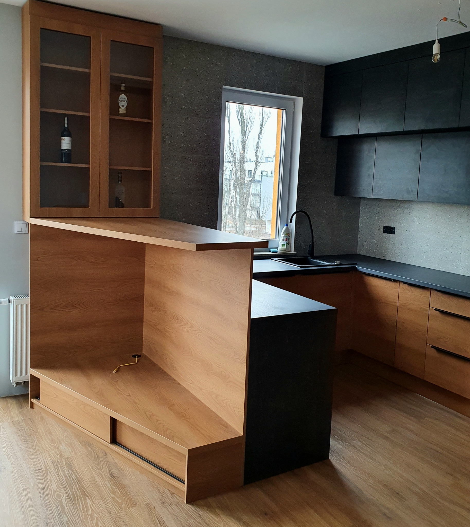 Nowoczesna kuchnia z drewnianymi szafkami, czarnymi blatami i barkiem, oświetlona wiszącą żarówką. Widoczny fragment okna z widokiem na budynek za drzewami bez liści.