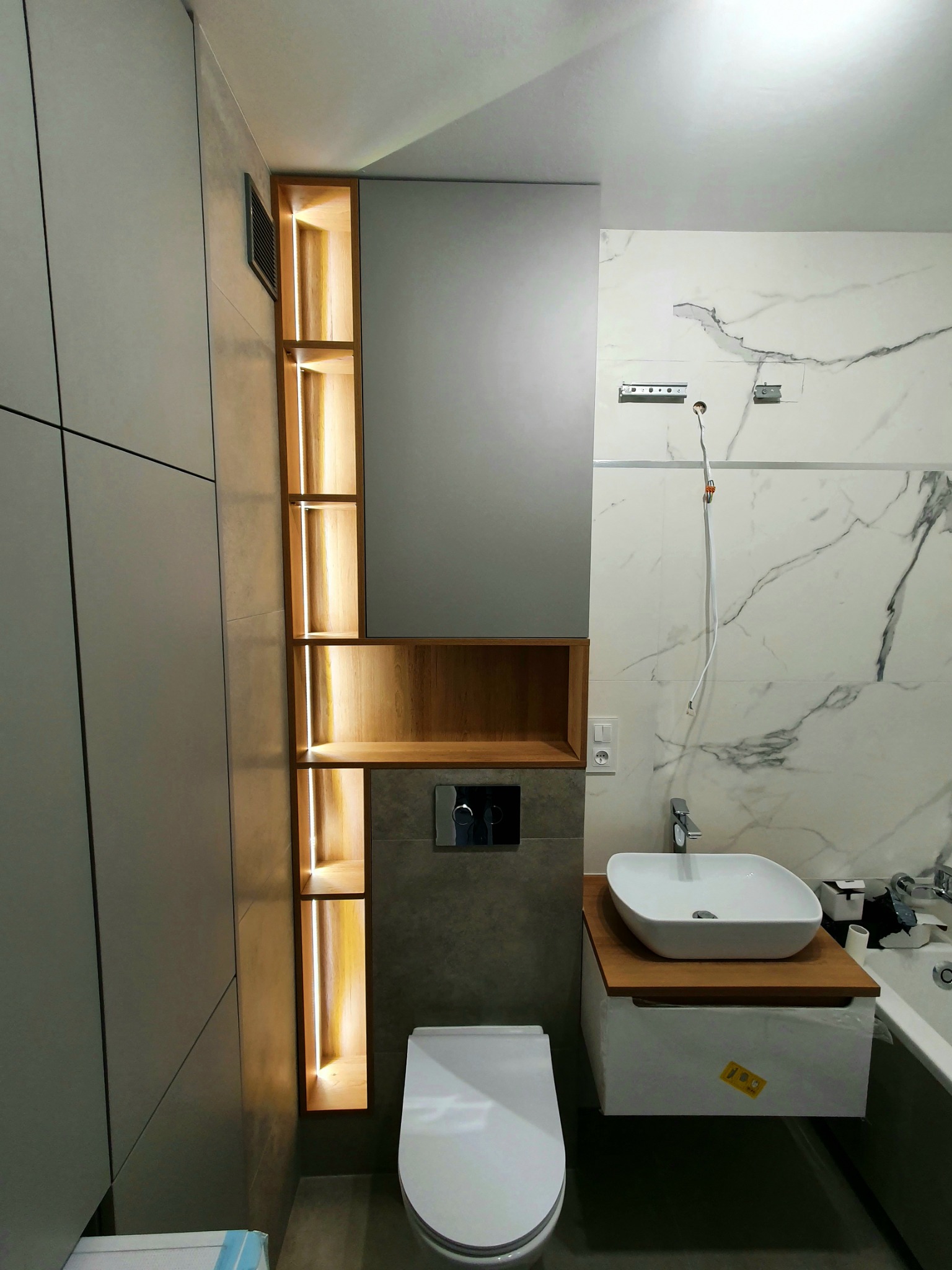 Nowoczesna łazienka w trakcie wykańczania: szara zabudowa meblowa z podświetlanymi półkami z drewna, wisząca toaleta, umywalka nablatowa na drewnianym blacie, marmurowe płytki częściowo zamontowane.