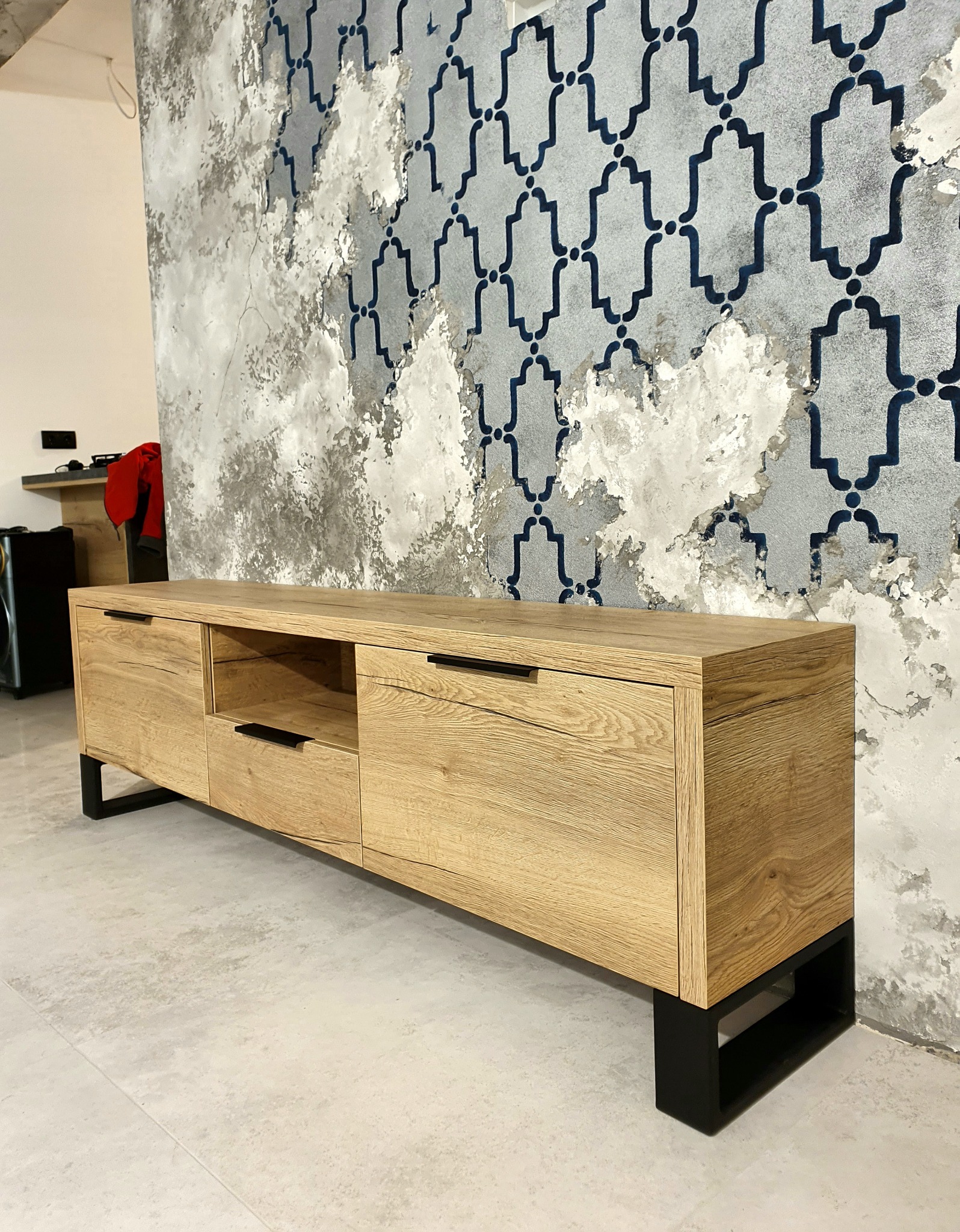 Drewniana szafka RTV z czarnymi uchwytami i nogami, na tle ściany z dekoracyjnym wzorem w odcieniach szarości i błękitu.