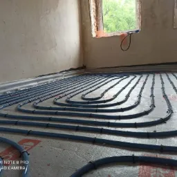 Niebieskie rury ogrzewania podłogowego ułożone serpentyną na izolacji, widok z niskiej perspektywy w surowym wnętrzu z oknami.