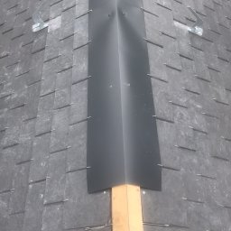 Usługi Stolarskie - Szary dach kryty łupkiem z zamontowaną, czarną obróbką blacharską na kalenicy, zabezpieczoną drewnianą łatą.