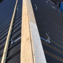 Czarny dach z blachy trapezowej w trakcie montażu, widoczne drewniane łaty i metalowe uchwyty śniegowe, rusztowanie w tle.