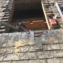 Usługi Stolarskie - Fragment dachu z szarej dachówki łupkowej, z wyciętym otworem, obok którego leży piła łańcuchowa i pudełko, widoczne drewniane elementy konstrukcji i trociny.