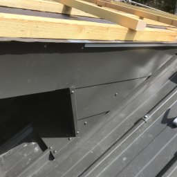 Usługi Stolarskie - Szary metalowy element obróbki blacharskiej na dachu z widoczną konstrukcją drewnianą w tle, na etapie montażu.