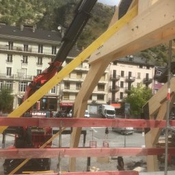 Usługi Stolarskie - Montaż drewnianej konstrukcji dachowej z użyciem dźwigu na tle zabudowy miejskiej i górskiego krajobrazu.