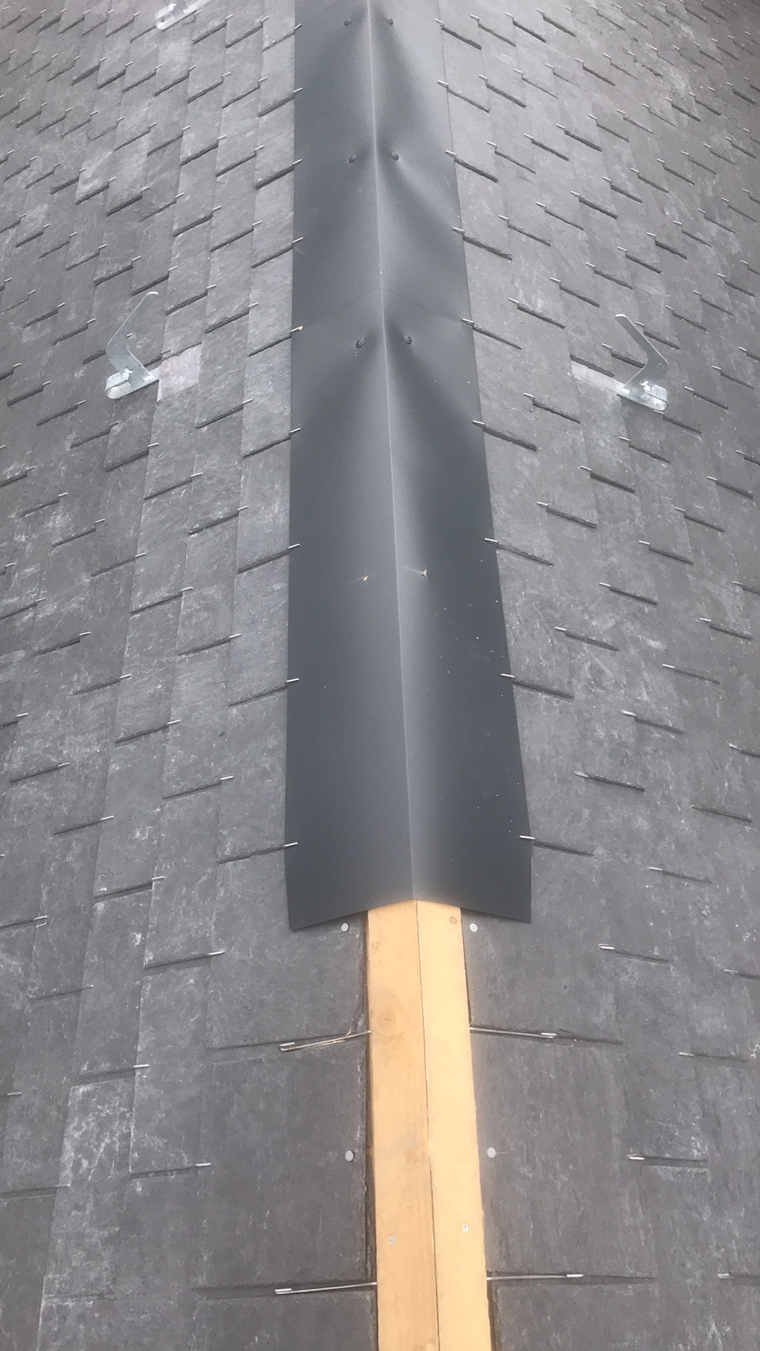 Szary dach kryty łupkiem z zamontowaną, czarną obróbką blacharską na kalenicy, zabezpieczoną drewnianą łatą.