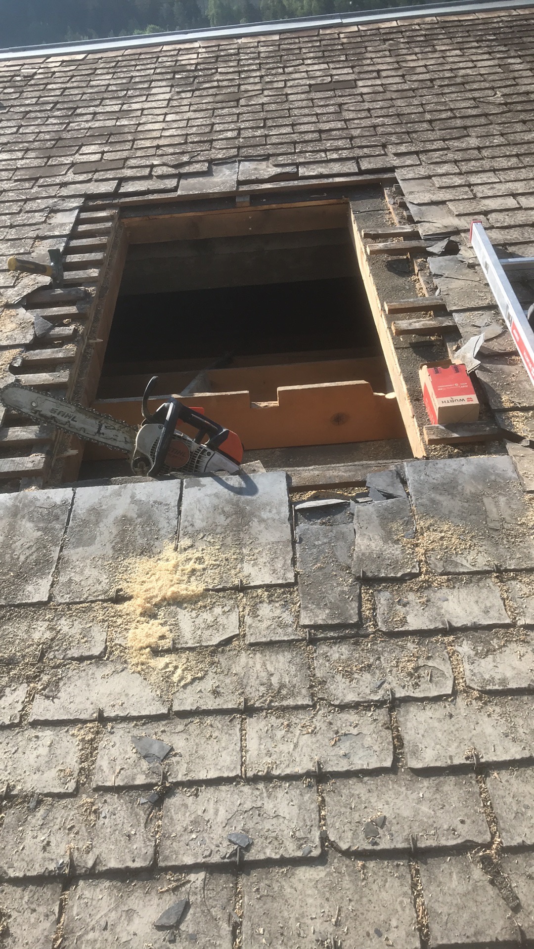 Fragment dachu z szarej dachówki łupkowej, z wyciętym otworem, obok którego leży piła łańcuchowa i pudełko, widoczne drewniane elementy konstrukcji i trociny.