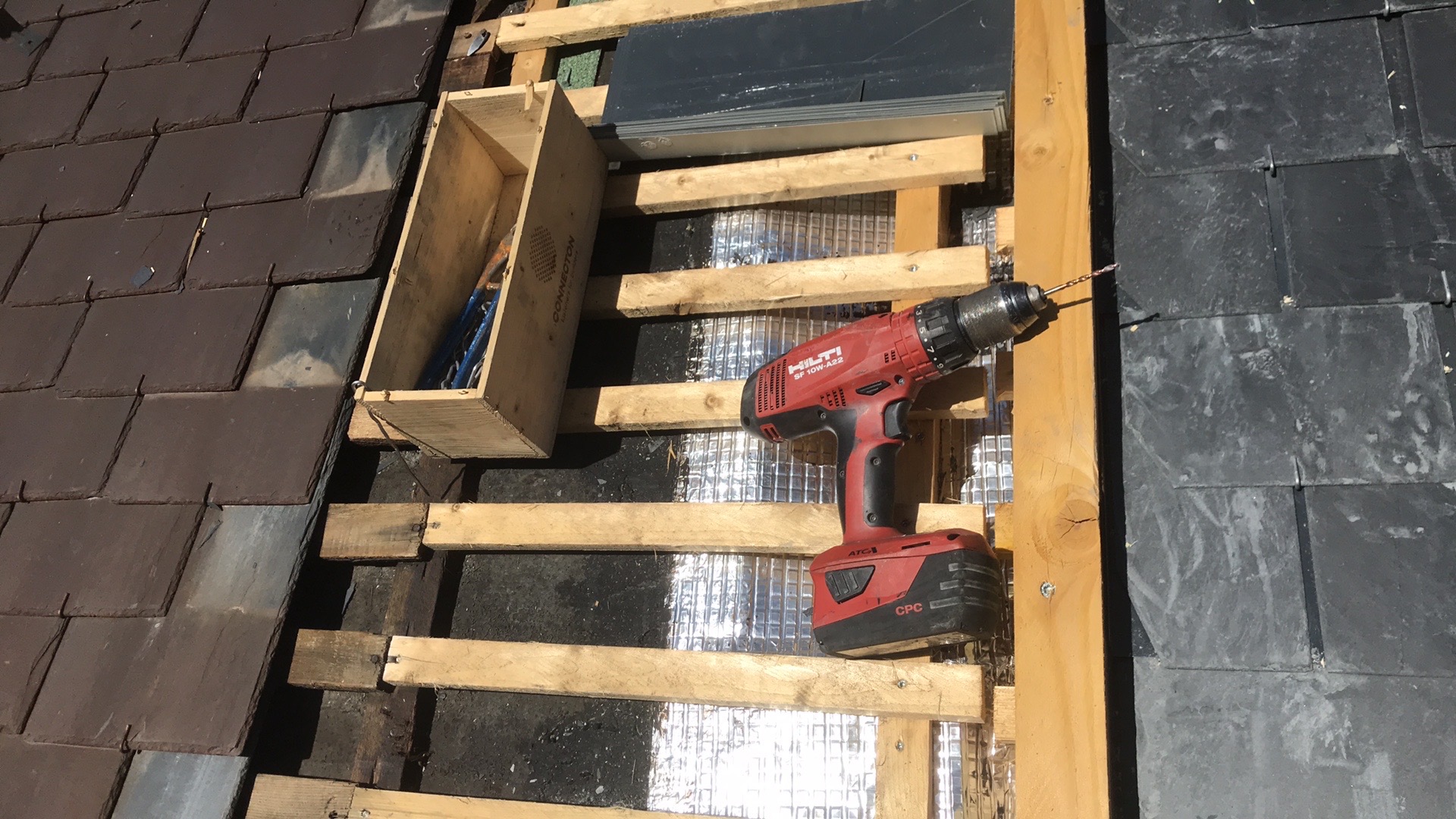 Częściowo zdjęte pokrycie dachu z łupka, widoczna drewniana konstrukcja, izolacja, skrzynka z narzędziami i wkrętarka Hilti.