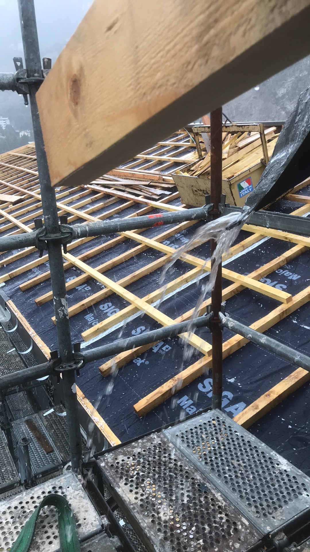 Konstrukcja dachu w trakcie budowy, widok z rusztowania w deszczowy dzień, drewniane łaty na czarnej membranie, woda spływająca po metalowych elementach rusztowania.