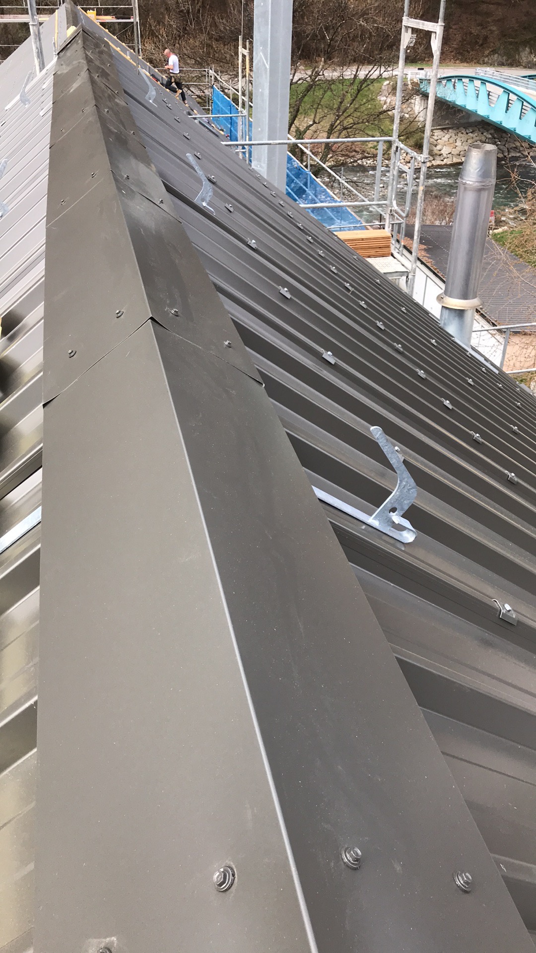 Nowy, metalowy dach w kolorze grafitowym z widocznymi elementami mocującymi i zabezpieczeniami śniegowymi, na tle rusztowania i mostu.