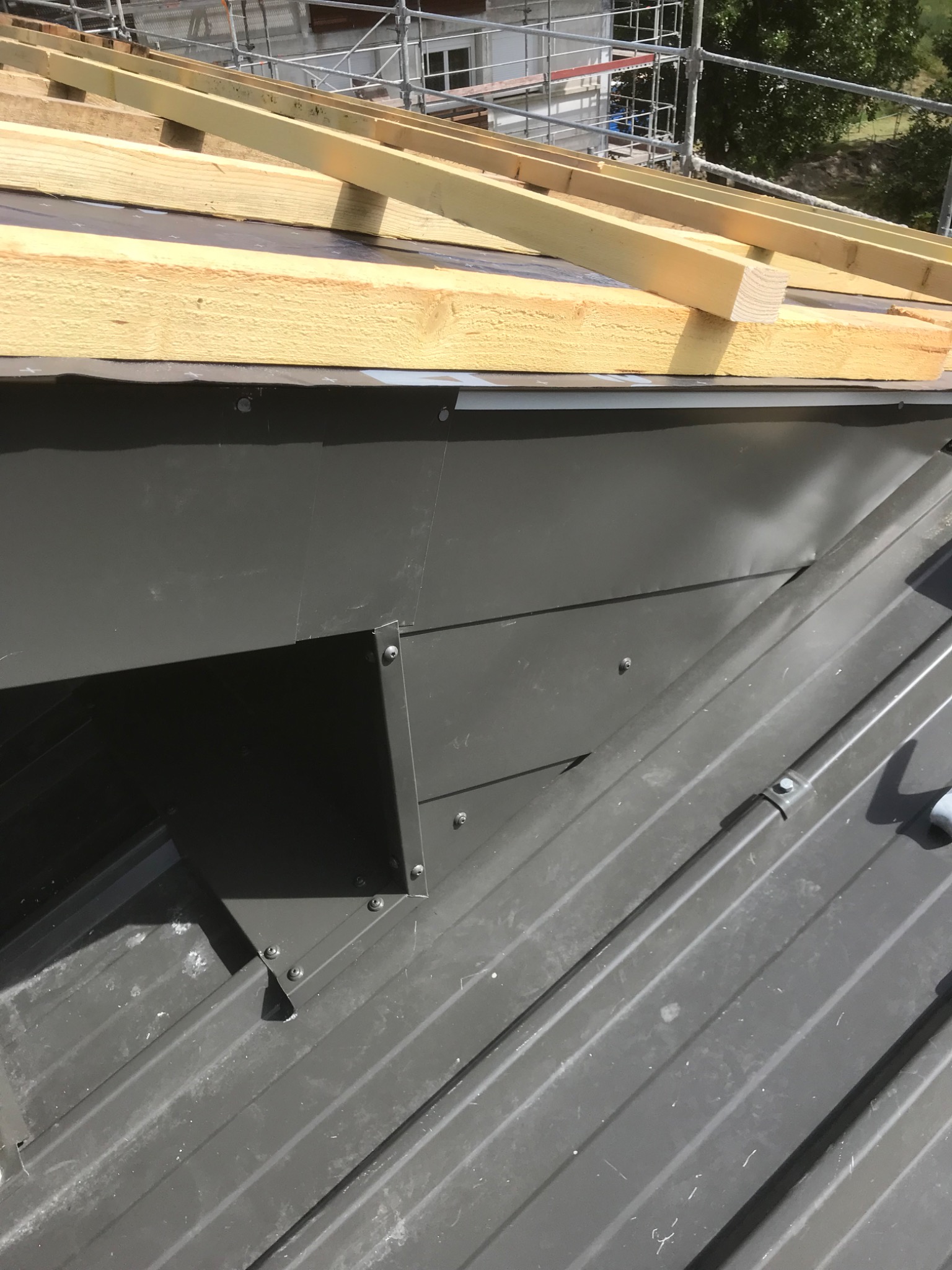 Szary metalowy element obróbki blacharskiej na dachu z widoczną konstrukcją drewnianą w tle, na etapie montażu.