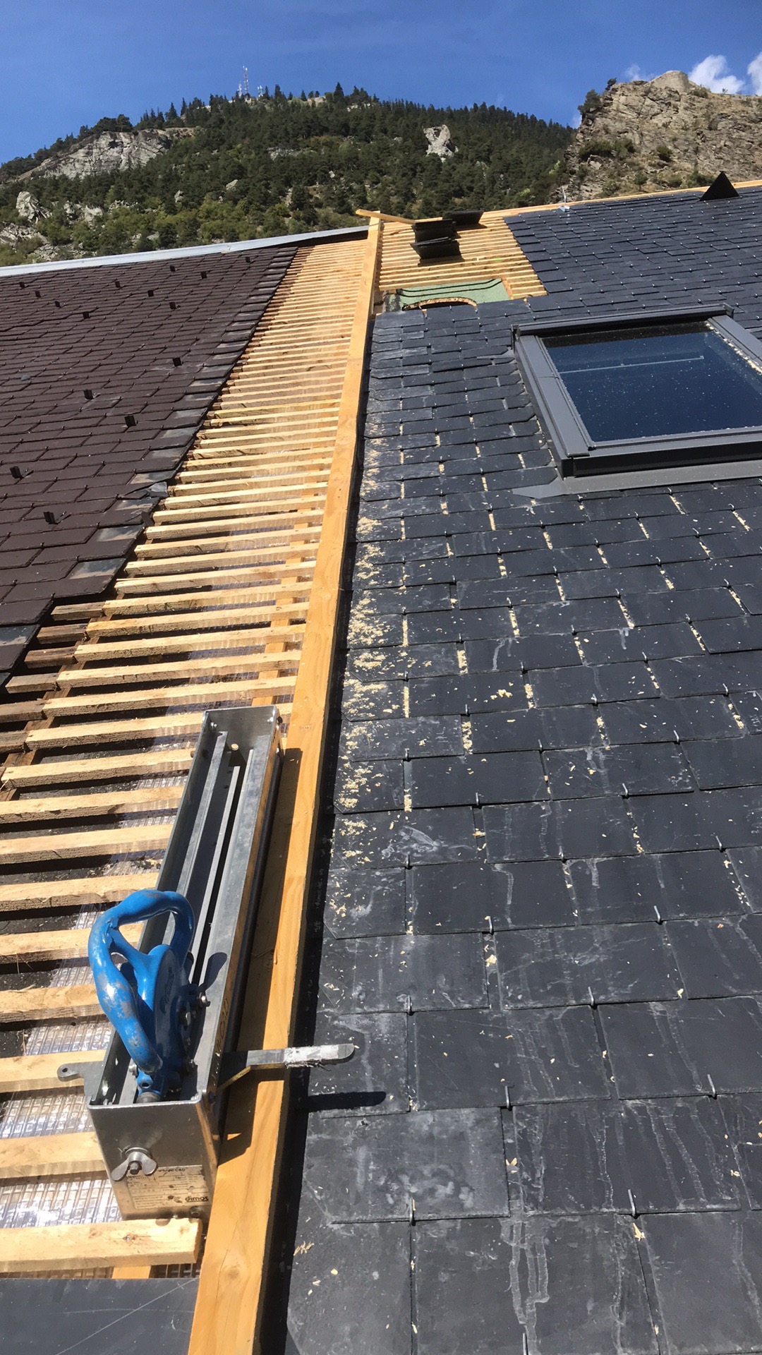 Fragment dachu w trakcie prac: część zdemontowana z widoczną konstrukcją drewnianą, część pokryta ciemnymi płytkami, okno dachowe, na tle górski krajobraz.