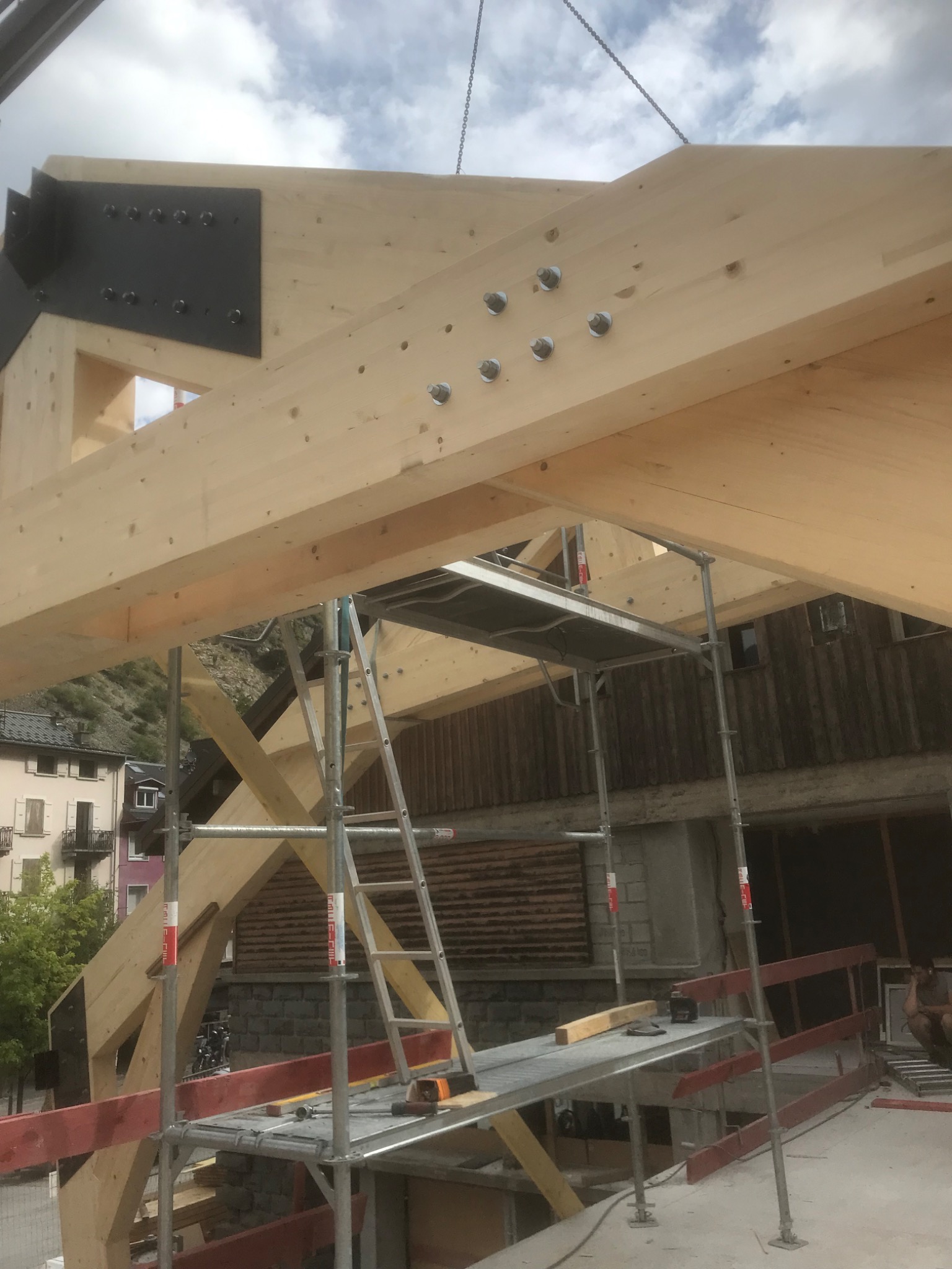 Montaż drewnianej konstrukcji dachu z użyciem metalowych łączników, widoczne rusztowania i otoczenie zabudowy mieszkalnej.