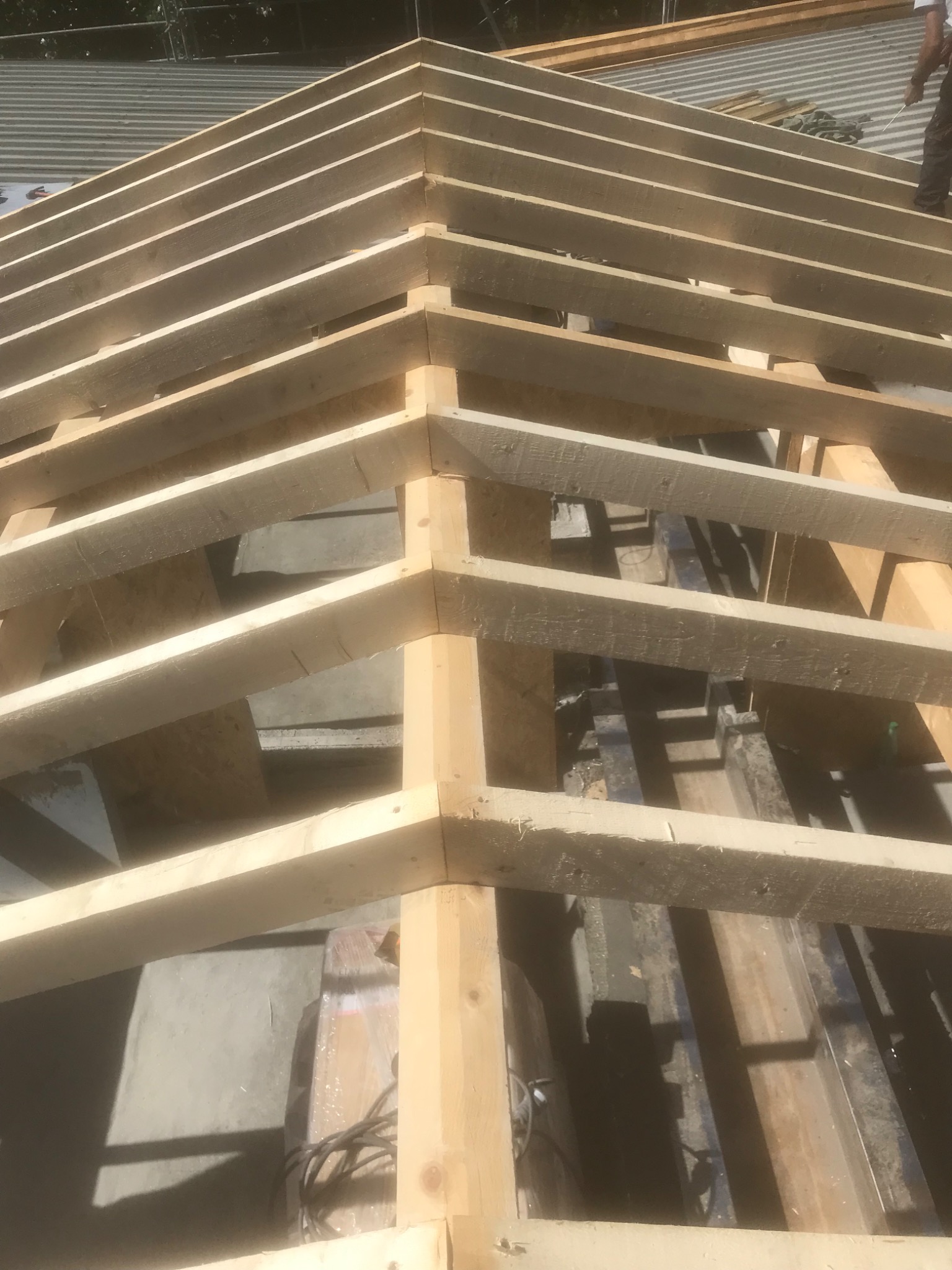 Konstrukcja drewniana więźby dachowej z widocznym fragmentem pokrycia dachowego w tle, częściowo oświetlona słońcem.