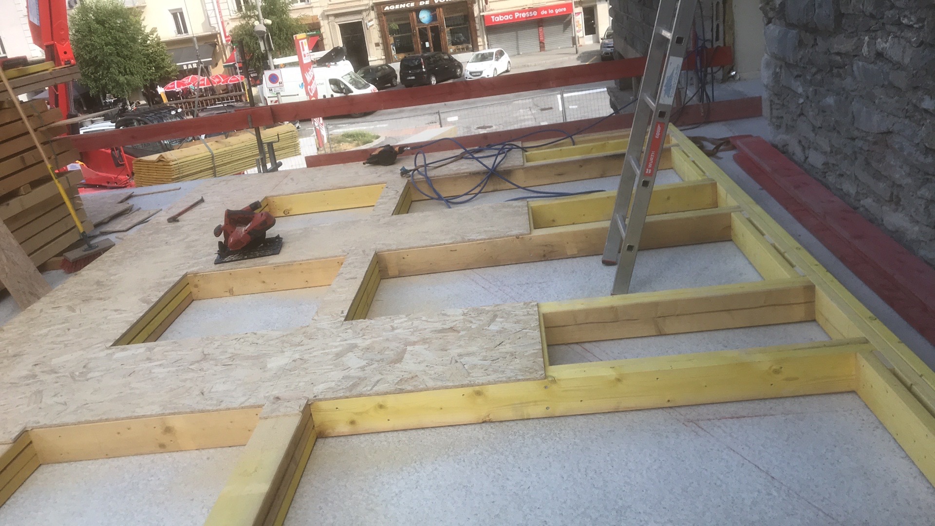 Konstrukcja dachu w trakcie budowy z widoczną deską OSB, żółtymi belkami, narzędziami i drabiną na tle miejskiej zabudowy.