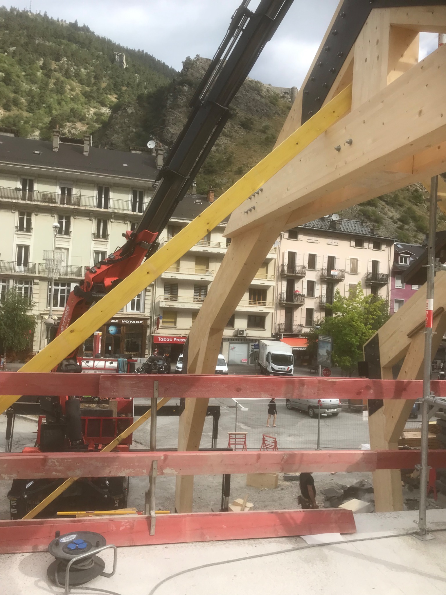 Montaż drewnianej konstrukcji dachowej z użyciem dźwigu na tle zabudowy miejskiej i górskiego krajobrazu.