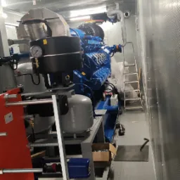 Wnętrze kontenera z zainstalowanym, niebieskim silnikiem spalinowym z osprzętem, widoczne manometry, rury, drabina i skrzynka z narzędziami.