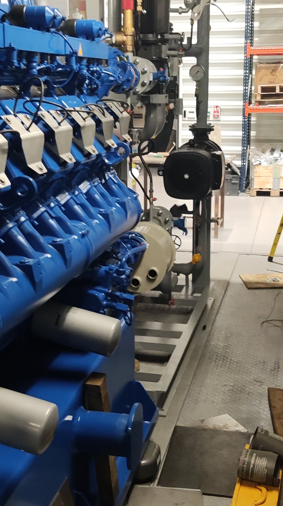 Zdjęcie przedstawia fragment dużej, przemysłowej instalacji grzewczej z niebieskimi elementami, widoczne liczne rury, zawory i zbiorniki, na tle magazynu z metalowymi regałami.