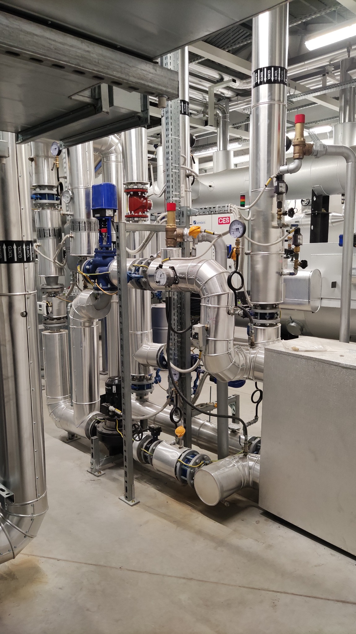 Kompleksowa instalacja rur z zaworami, manometrami i izolacją termiczną w pomieszczeniu technicznym, widoczne rury z oznaczeniami GLYCOL.
