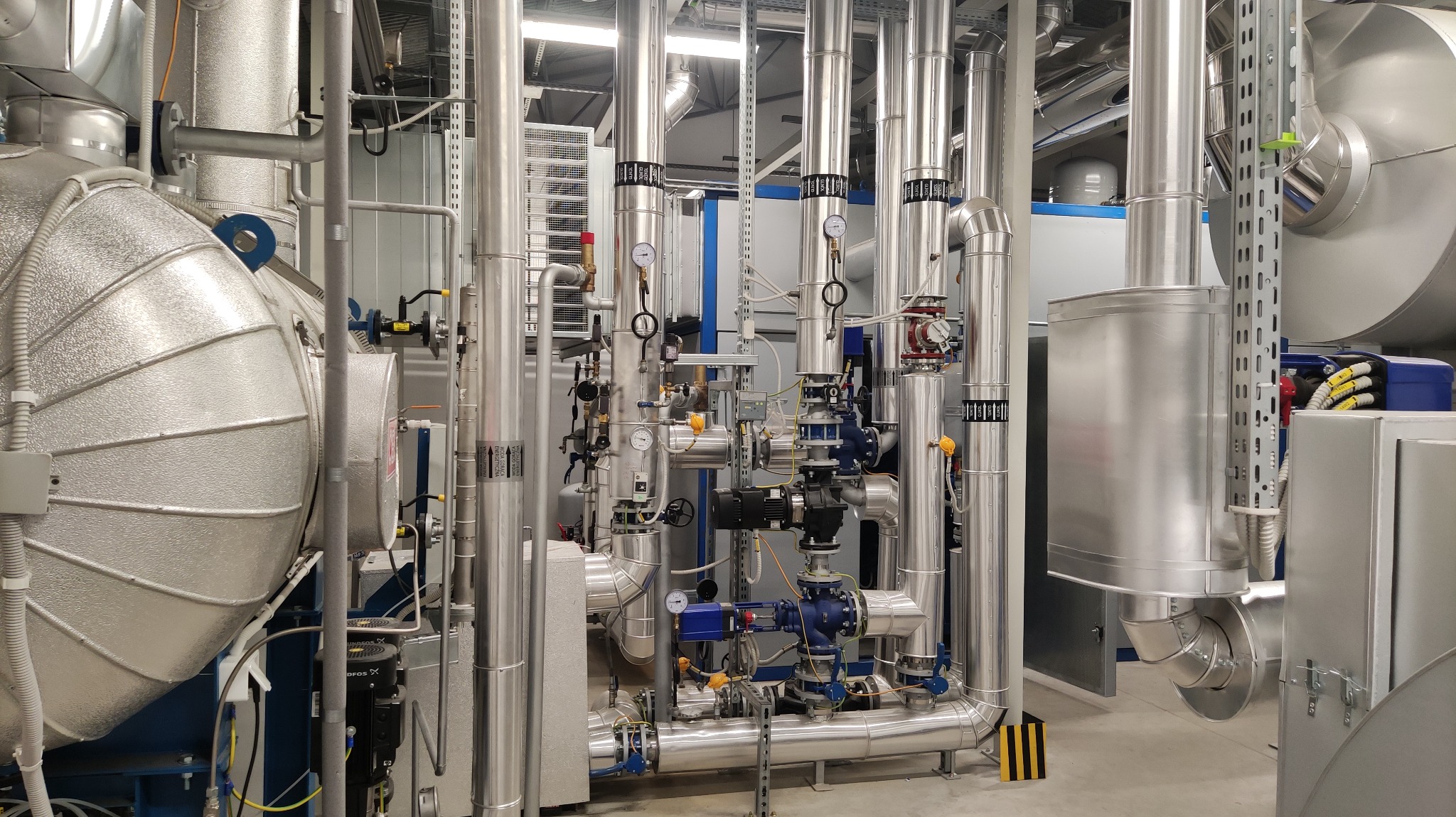 Rozbudowana instalacja przemysłowa z licznymi rurami, zaworami, manometrami i elementami izolacyjnymi, utrzymana w srebrnej kolorystyce, widoczna w hali z szarymi ścianami i podłogą.