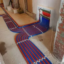 Rozdzielacz ogrzewania podłogowego z rurami PEX w kolorze czerwonym i niebieskim, zamocowanymi do ściany w surowym stanie.