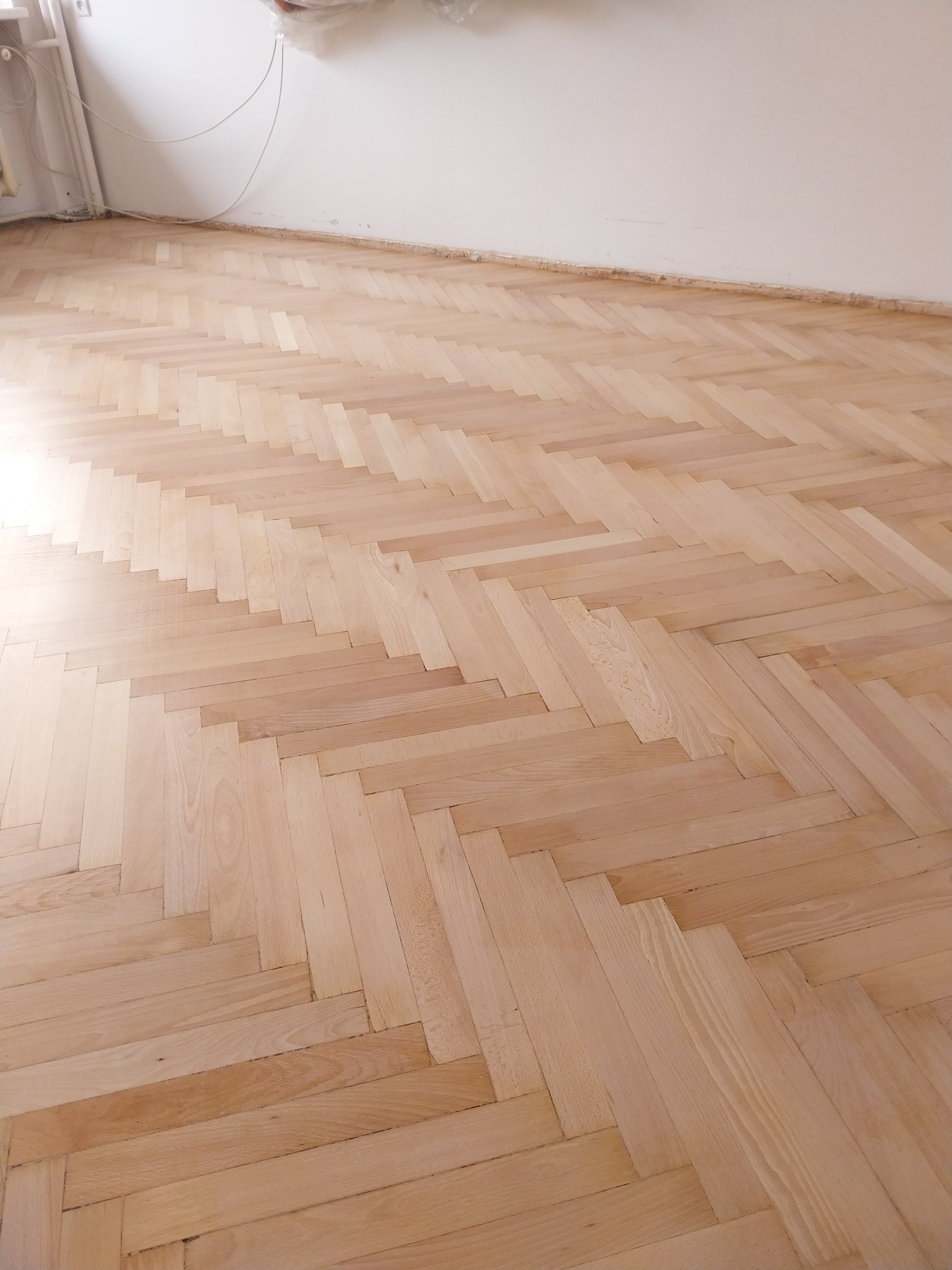 Jasny parkiet ułożony w jodełkę, świeżo po cyklinowaniu. Widoczne delikatne różnice w odcieniach drewna i minimalne niedoskonałości podkreślające naturalny charakter podłogi.