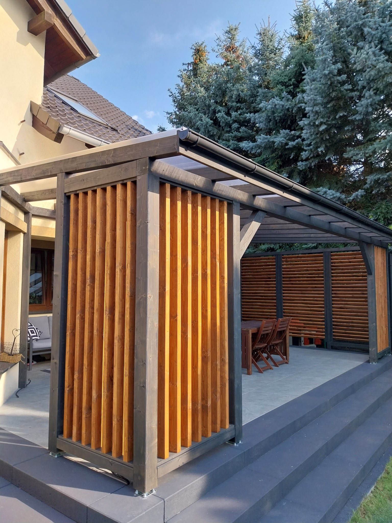 Drewniana pergola z lamelami w odcieniach brązu i szarości, zadaszona, z widocznym stołem i krzesłami, na tle zielonych drzew i fragmentu budynku.