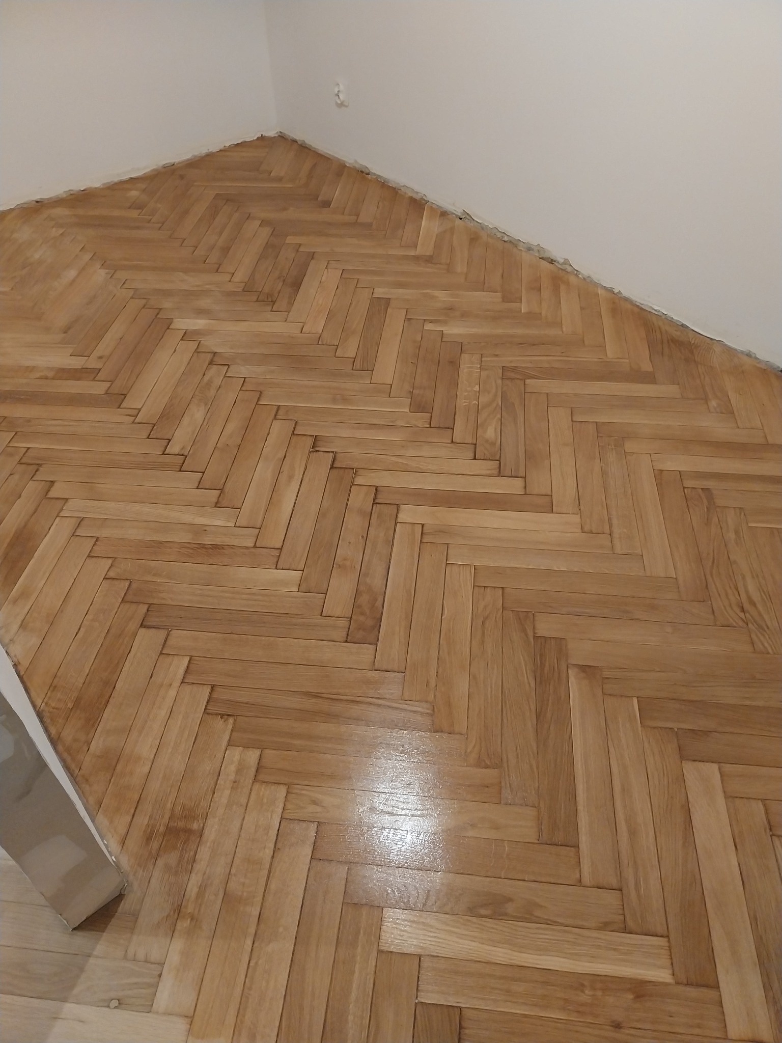 Ułożony parkiet w jodełkę, świeżo po cyklinowaniu, z widocznym połyskiem lakieru. Jasne drewno, minimalistyczne wnętrze, widoczny fragment listwy przypodłogowej.