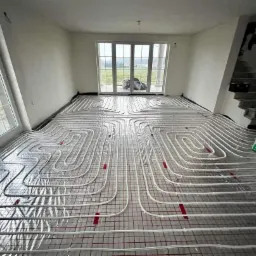 Białe rurki ogrzewania podłogowego ułożone na srebrnej folii z czerwoną siatką w surowym wnętrzu z dużymi oknami i widokiem na zewnątrz.