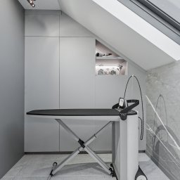 Adam Jasek Design Studio - Szare wnętrze ze skośnym sufitem, marmurową ścianą i deską do prasowania z generatorem pary; w tle zabudowa meblowa z otwartą półką.