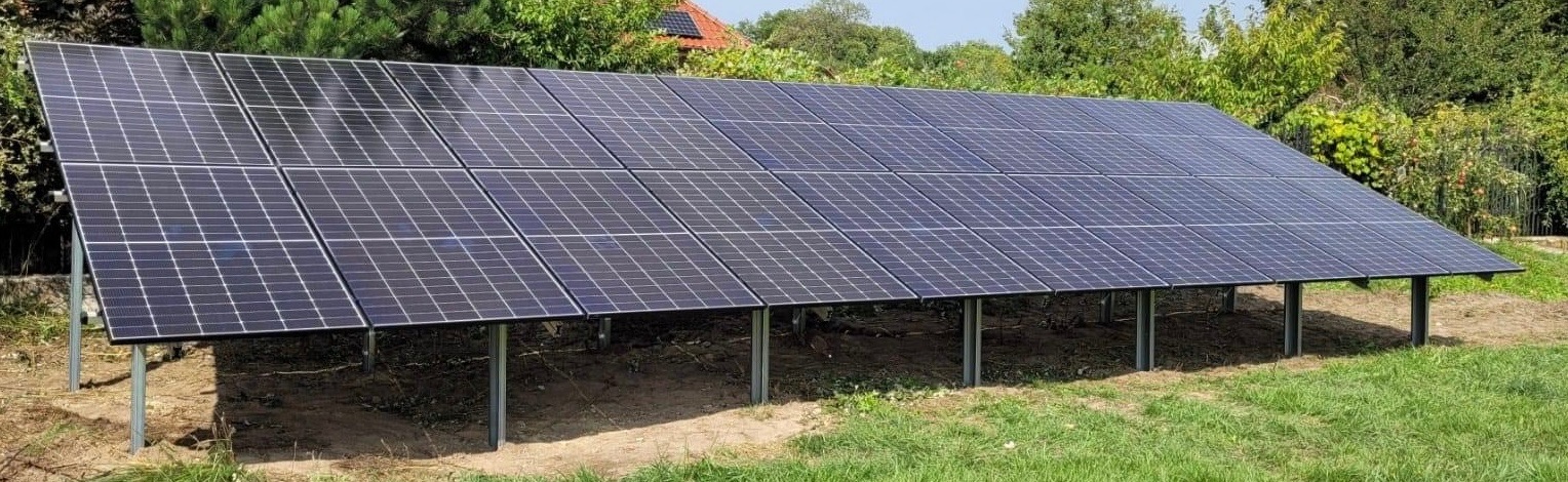 Instalacja paneli słonecznych na gruncie, widoczne metalowe podpory, trawa i drzewa w tle, na dachu domu również zamontowane panele.