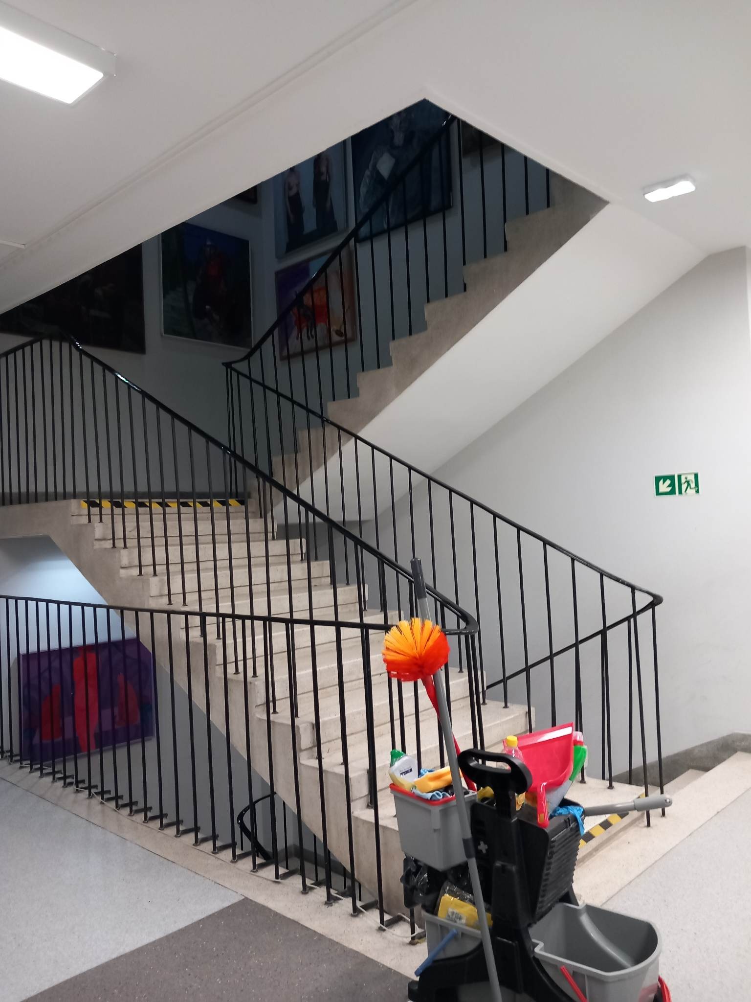Wózek do sprzątania z mopem i środkami czystości na schodach w biurowcu lub budynku użyteczności publicznej, widoczne obrazy na ścianie i żółto-czarne taśmy antypoślizgowe na stopniach.