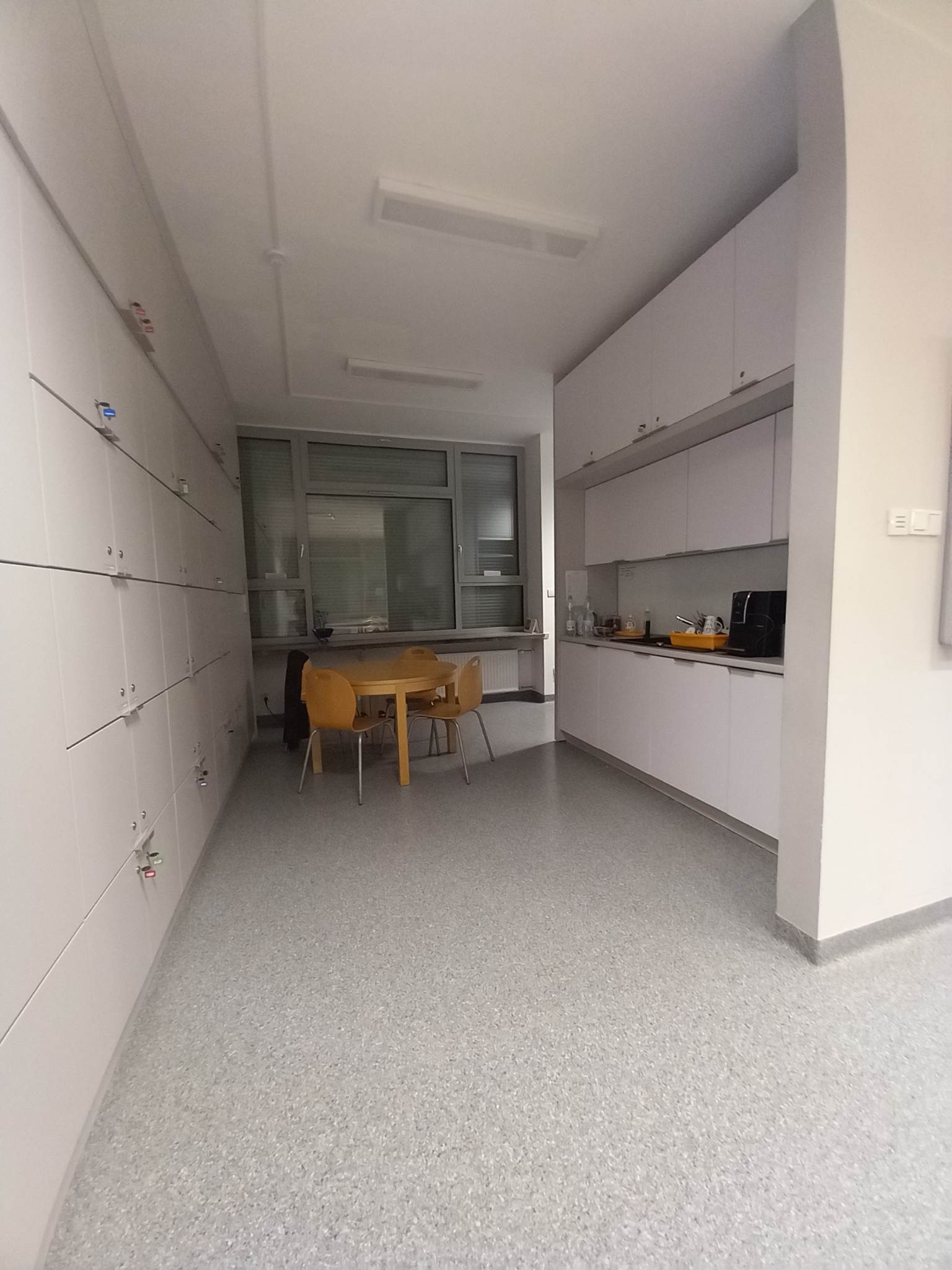 Wnętrze biura z rzędem białych szafek, stołem z krzesłami i aneksem kuchennym z białymi szafkami.