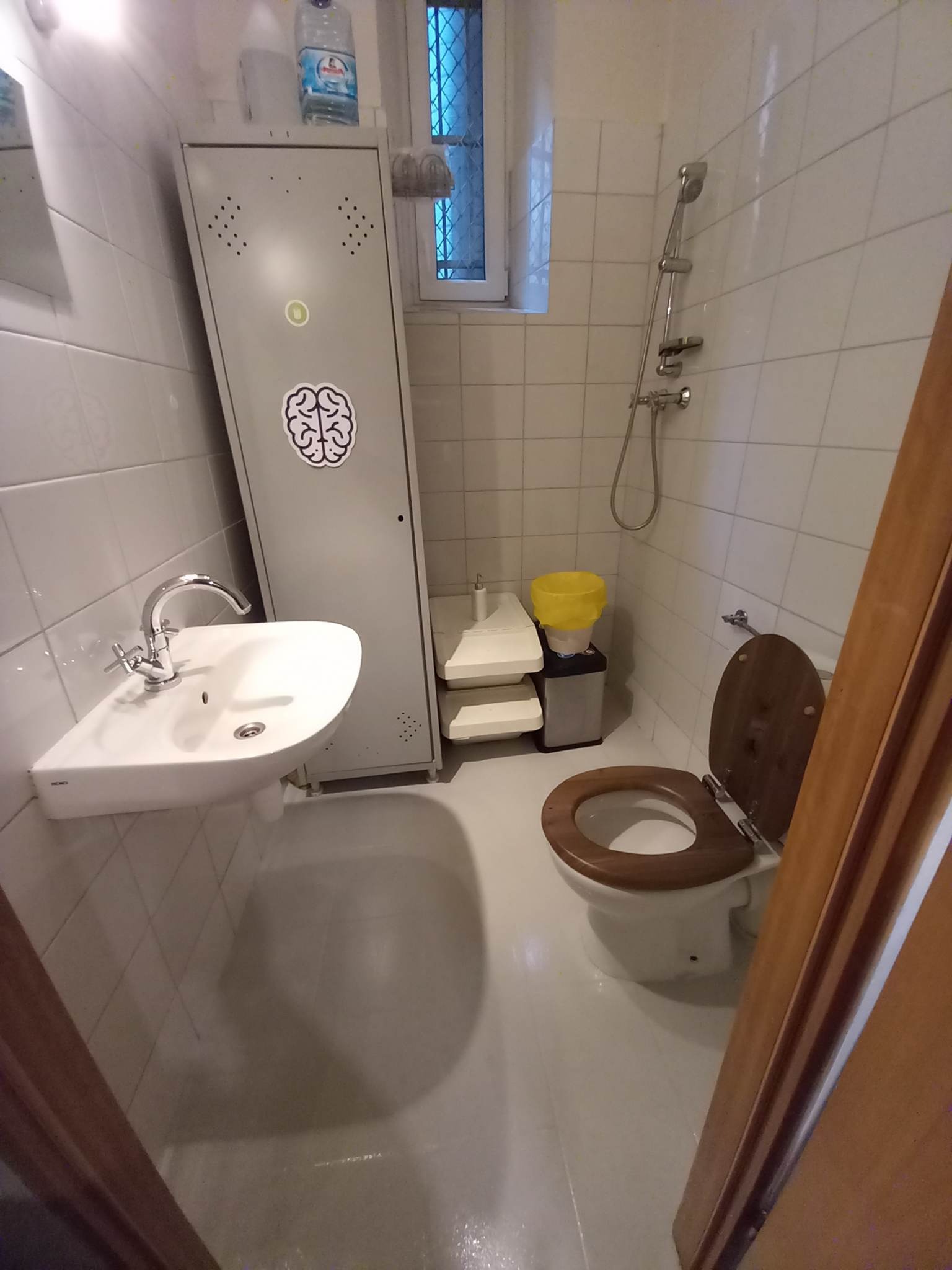 Wnętrze łazienki z umywalką, toaletą z drewnianą deską, prysznicem i metalową szafką ozdobioną naklejką z mózgiem.