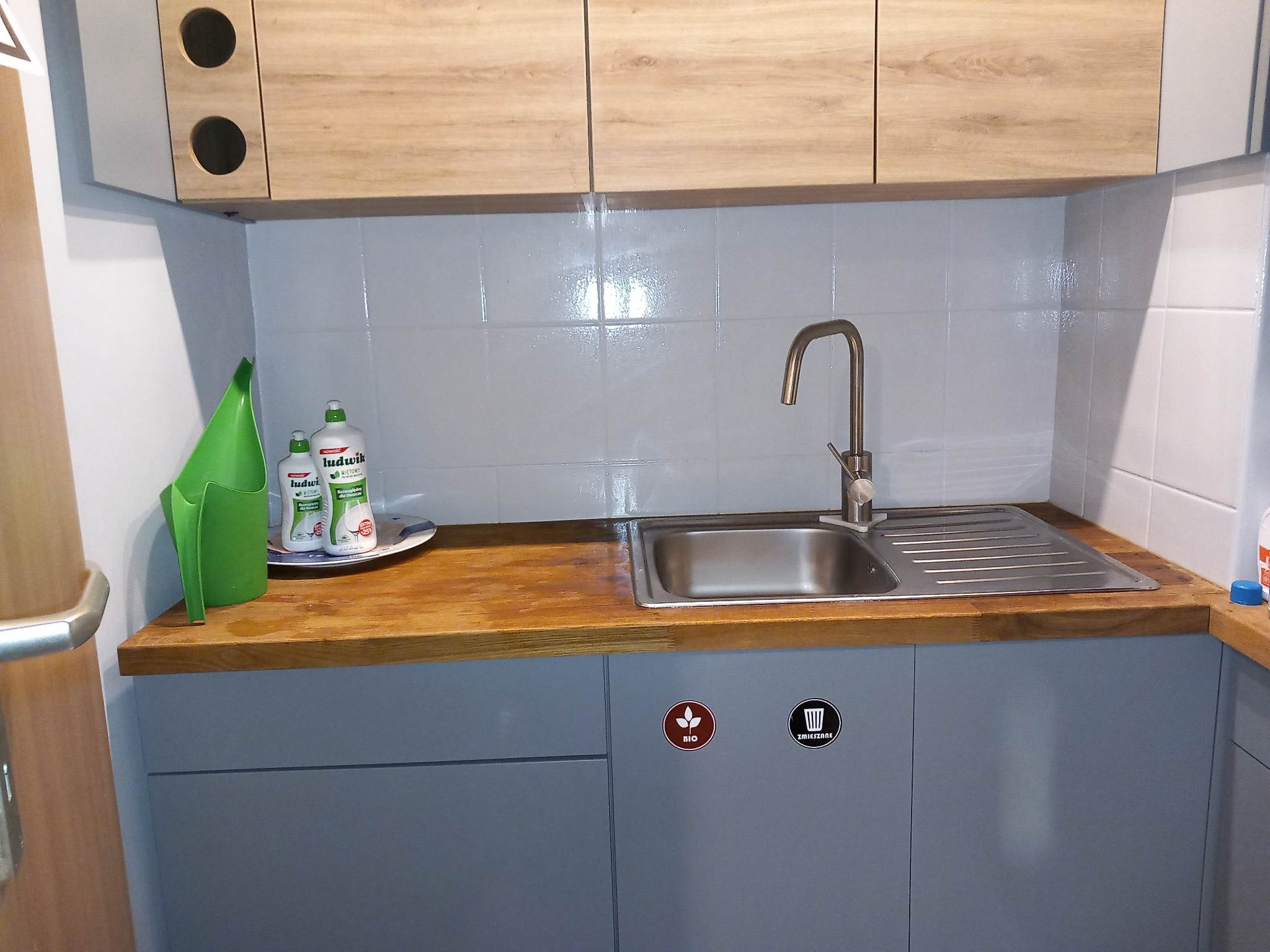 Umywalka ze stali nierdzewnej w blacie z drewna, nad szafkami z naklejkami do segregacji odpadów, z widocznymi środkami czystości Ludwik i zielonym dzbankiem.