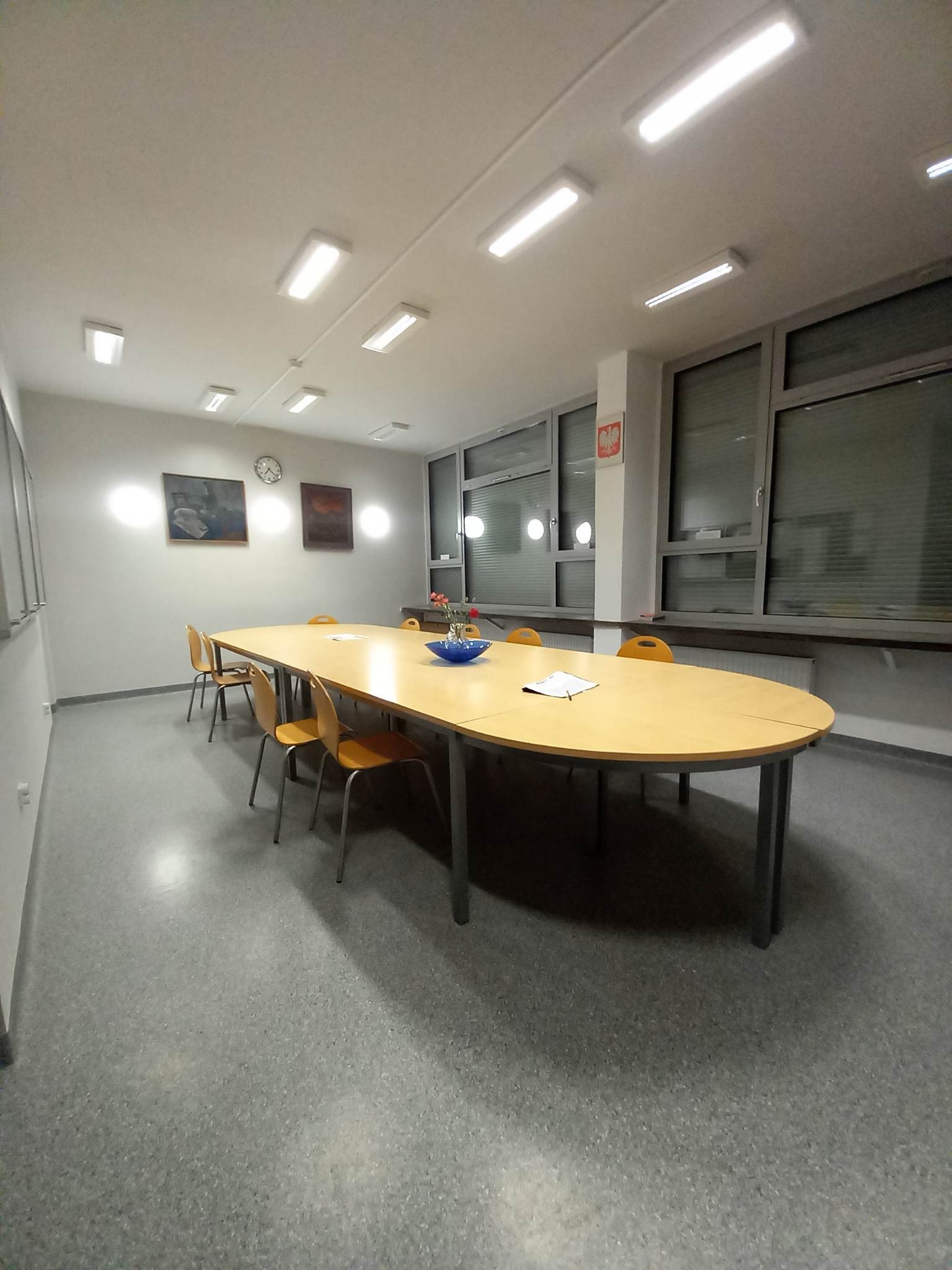 Sala konferencyjna z owalnym stołem, krzesłami, kwiatami w niebieskiej misie, obrazami na ścianie i polską flagą w tle, oświetlona jarzeniówkami.
