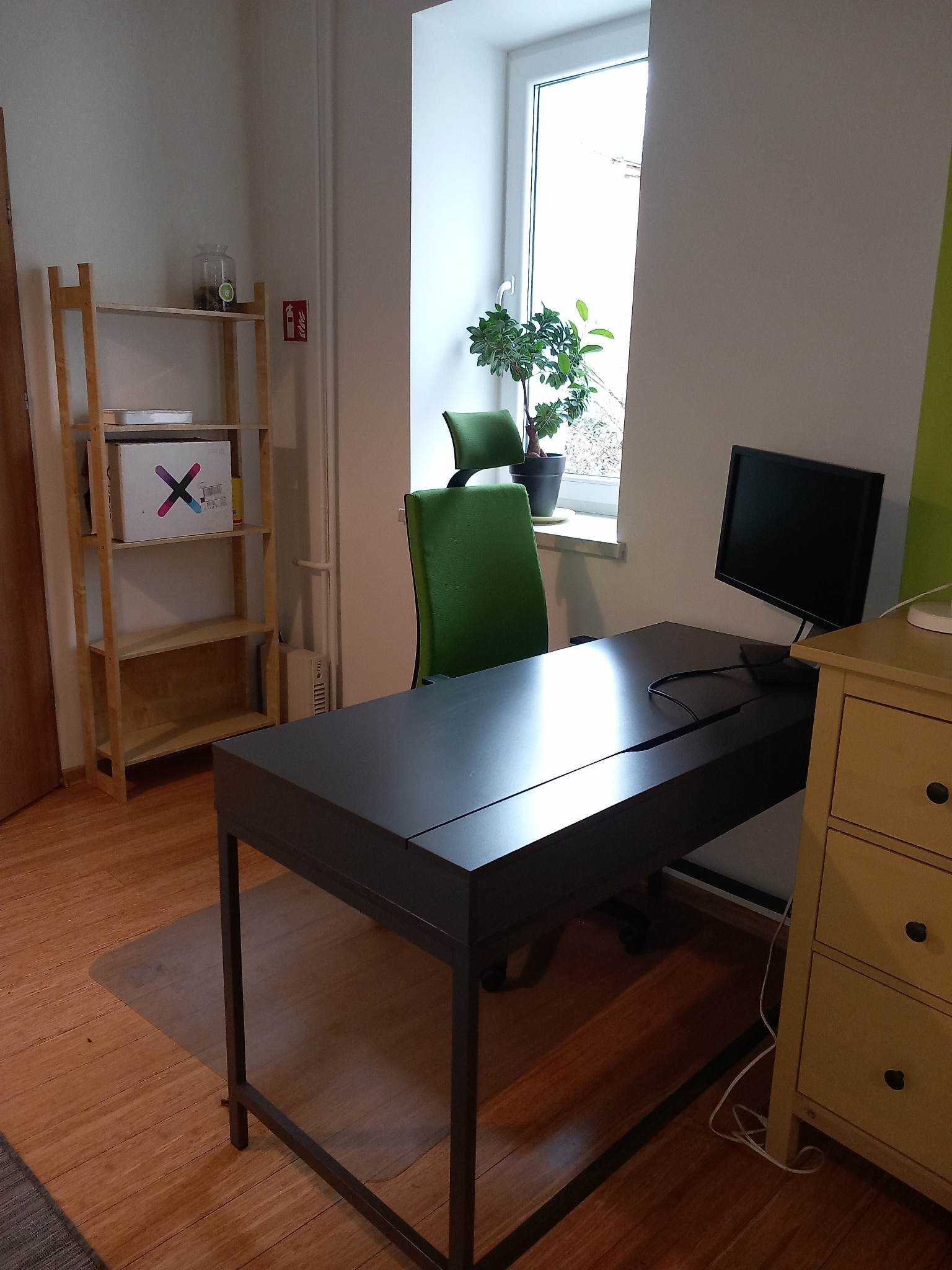 Nowoczesne biuro z ciemnym biurkiem, zielonym krzesłem i rośliną doniczkową na parapecie, uzupełnione drewnianym regałem i komodą w jasnym odcieniu.